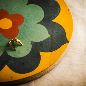 A small gold ring with a bird design, resting on an Athangudi Tile Tray with a retro floral pattern, set against a textured beige fabric background.