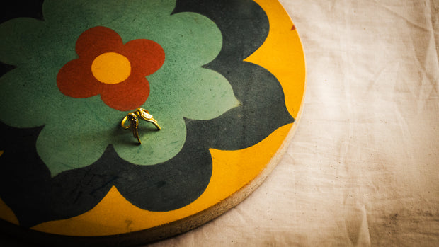 A small gold ring with a bird design, resting on an Athangudi Tile Tray with a retro floral pattern, set against a textured beige fabric background.